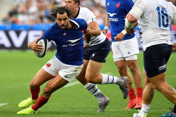 Un joueur en maillot bleu foncé porte le ballon tandis qu’un adversaire en maillot blanc tente de l’attraper sur un terrain de rugby