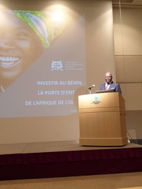 Un homme parle à un pupitre devant un grand écran affichant un message sur l'investissement au Bénin