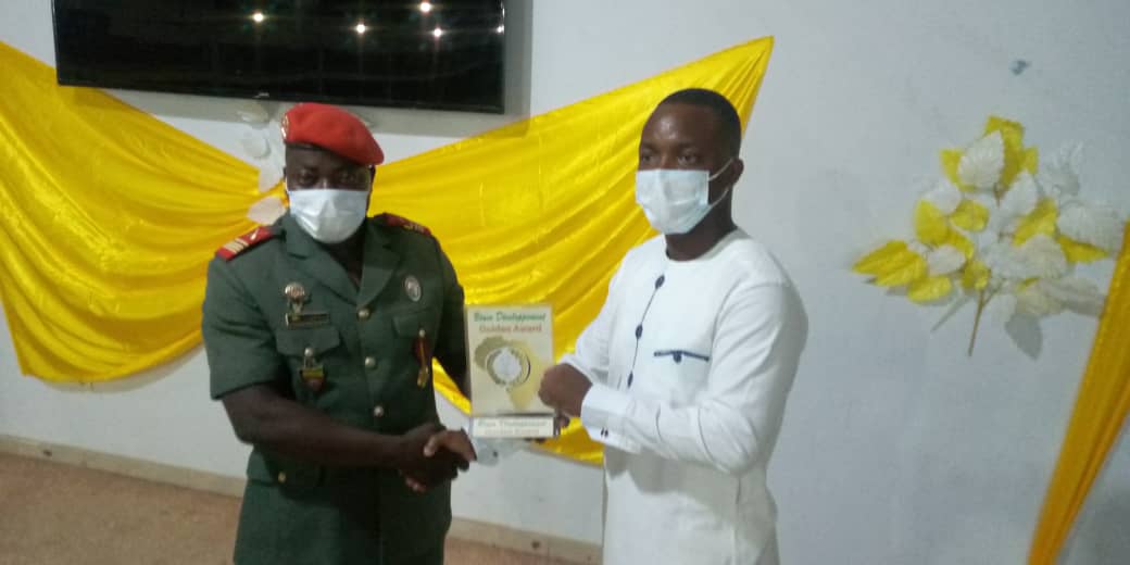 Deux hommes masqués s’échangent un trophée dans une pièce décorée de jaune avec un écran noir au mur