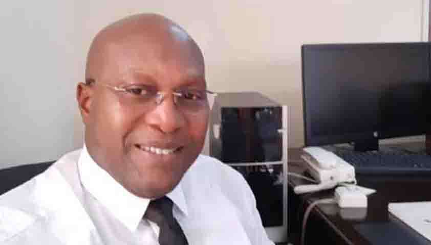 Homme portant une chemise blanche et une cravate noire souriant en bureau avec ordinateur et téléphone fixe visible en arrière-plan