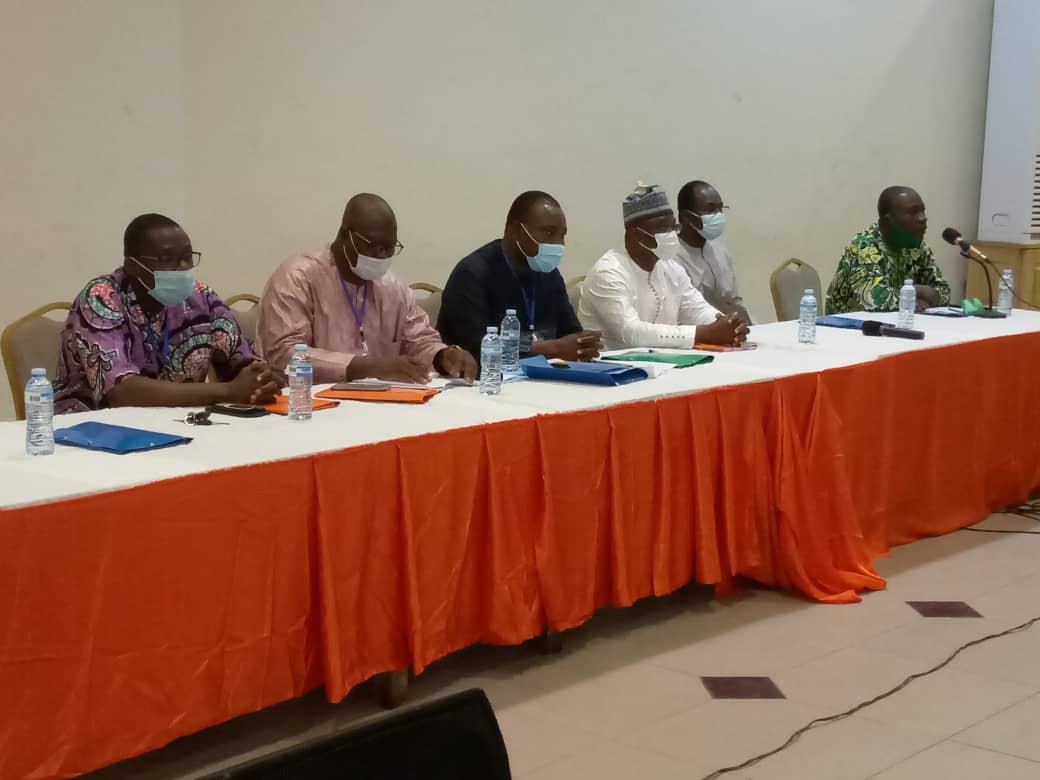 Six hommes assis à une longue table avec nappes orange et blanche portant des masques et préparant des documents