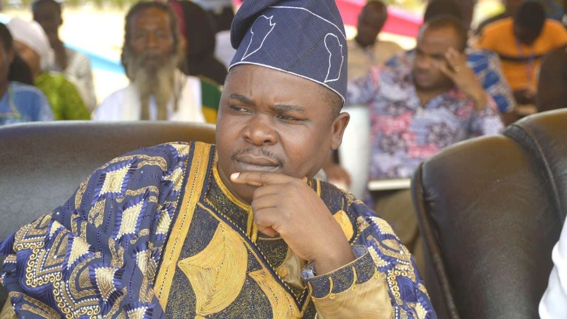 Un homme en tenue traditionnelle africaine regarde pensivement sur le côté entouré de plusieurs personnes floues en arrière-plan