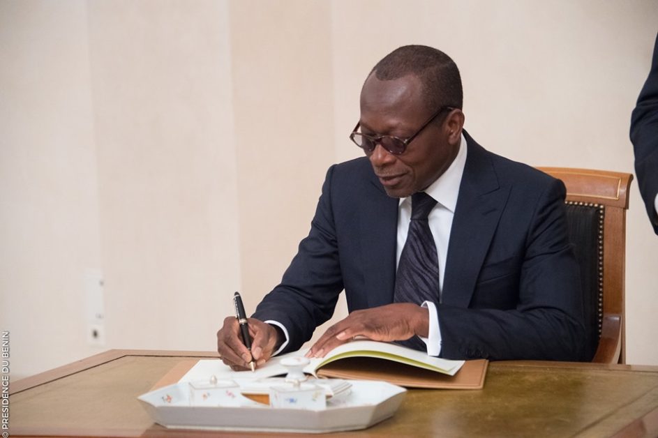 Un homme en costume sombre signe un document assis à une table avec un plateau blanc devant lui
