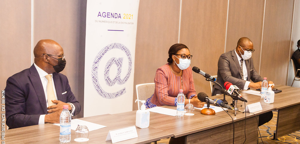 Trois personnes masquées assises à une table lors d'une conférence avec un panneau "Agenda 2021" en arrière-plan