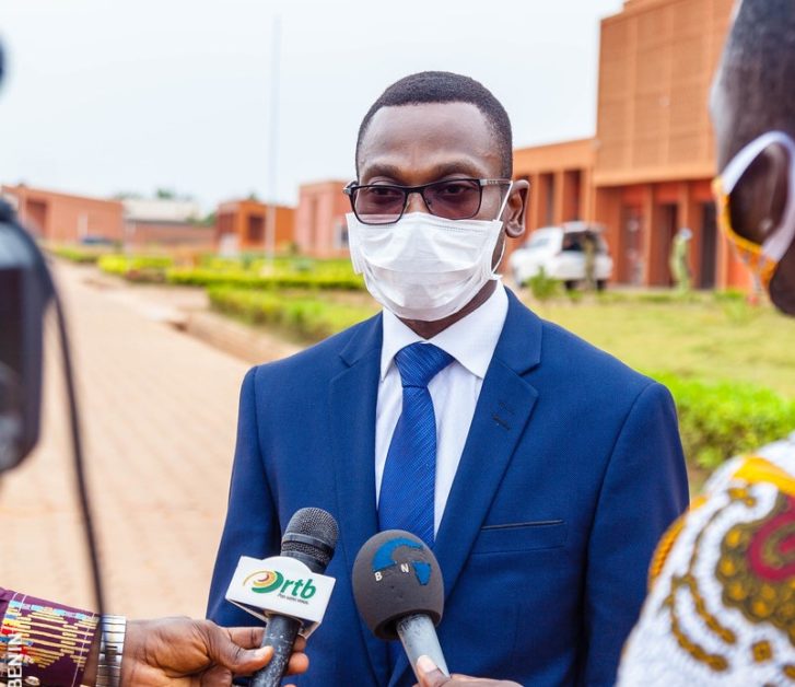Homme en costume bleu et cravate bleue portant un masque blanc répond à des journalistes avec micros devant un bâtiment rouge.