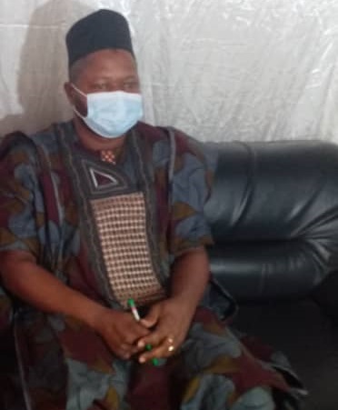 Homme portant un boubou traditionnel et un masque assis sur un canapé noir les mains jointes.