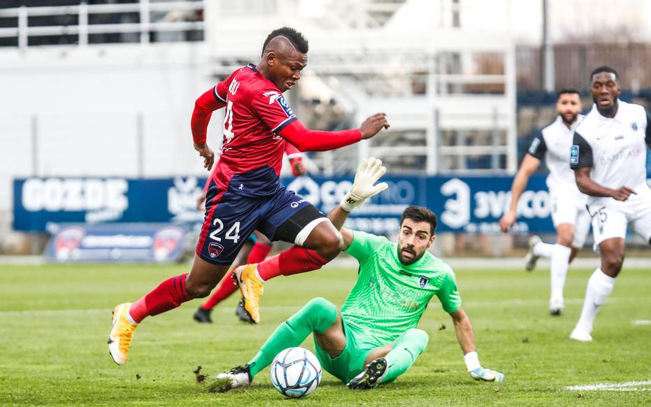 Un joueur en maillot rouge saute pour éviter le gardien en vert qui tente d’intercepter le ballon sur un terrain extérieur avec plusieurs joueurs en arrière-pla