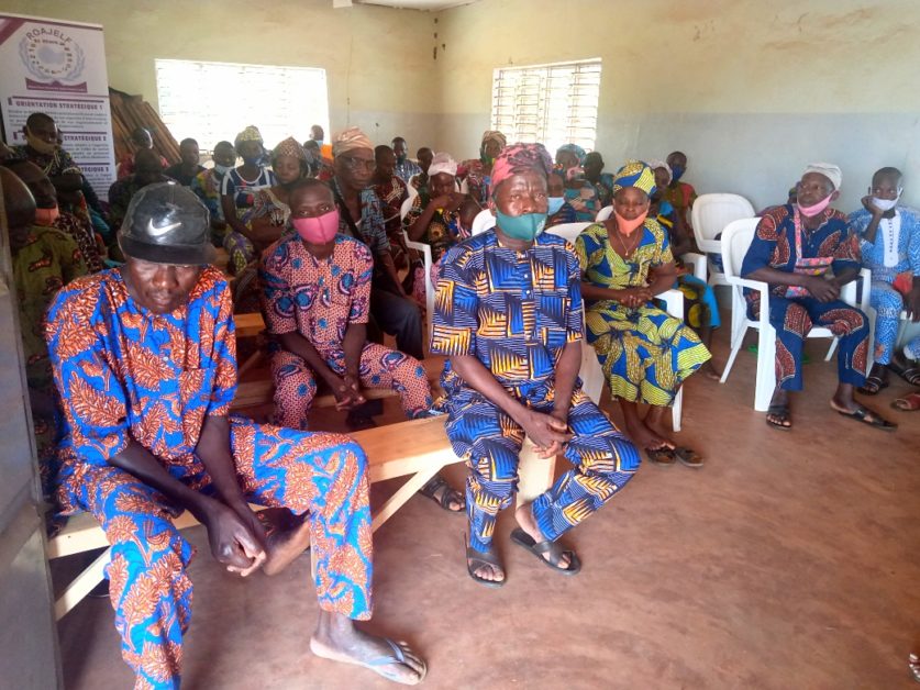 Groupe d’hommes et de femmes en pagnes traditionnels assis dans une salle aux murs jaunes et bancs en bois et chaises blanches