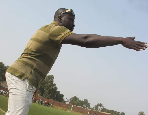 Un homme en polo rayé vert et blanc fait un geste de bras tendu sur un terrain de football en extérieur