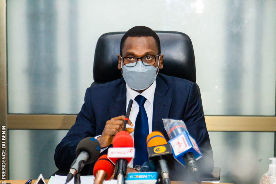 Homme en costume et masque parlant devant plusieurs micros lors d'une conférence de presse assis à un bureau