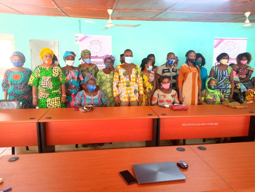 Un groupe de femmes habillées de façon colorée portant des masques posent debout derrière une table avec un ordinateur portable posé dessus