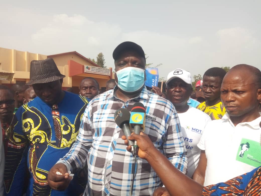Un homme avec masque et casquette parle face à plusieurs micros devant un groupe d'hommes lors d'un rassemblement en plein air