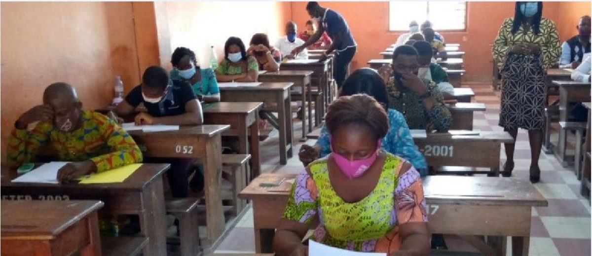 Des candidats masqués passent un examen assis individuellement à des pupitres dans une salle de classe éclairée par la lumière du jour