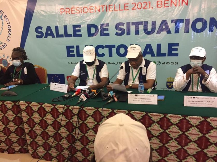 Quatre personnes assises à une table avec des micros et un grand panneau « Salle de situation électorale » en arrière-plan