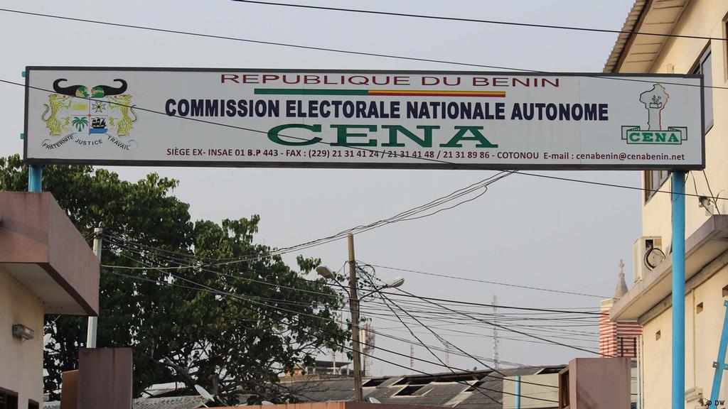 Panneau suspendu avec texte « République du Bénin » et « Commission Électorale Nationale Autonome CENA » au-dessus d’une rue urbaine avec fils électriques visib