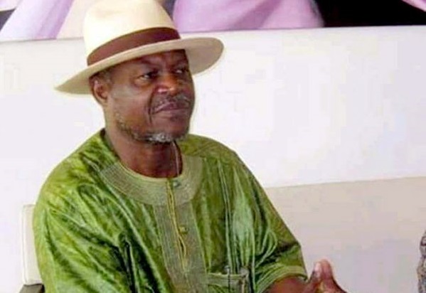 Homme portant un chapeau clair et une tenue verte assis, regardant devant lui avec les mains jointes