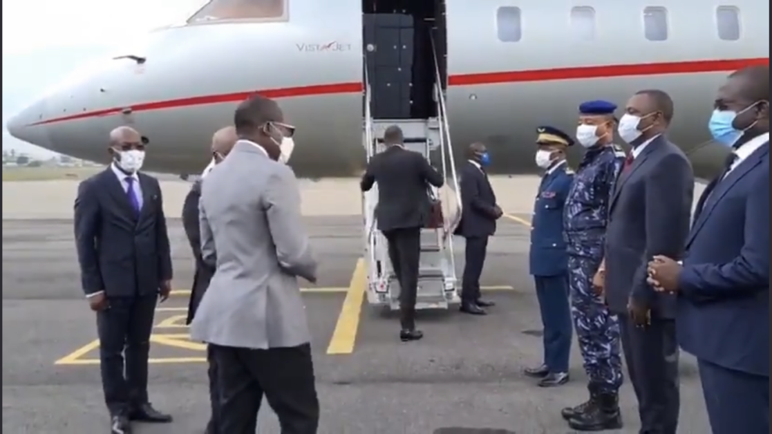 Homme en costume montant les marches d’un avion privé entouré de plusieurs personnes en costume et uniforme portant un masque