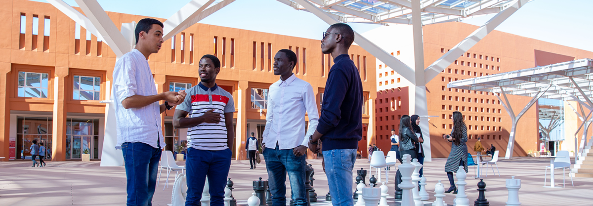 Quatre jeunes hommes discutent autour d’un jeu d’échecs géant en plein air dans une cour moderne ensoleillée