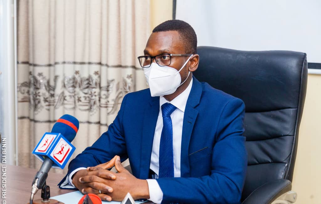 Homme en costume bleu et cravate blanche portant un masque assis derrière un bureau avec microphones devant lui