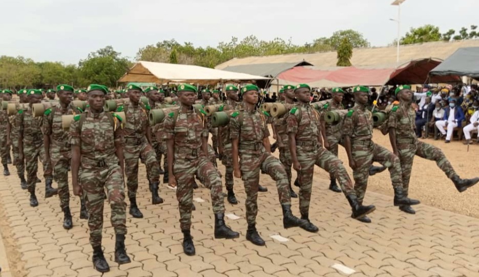 Des militaires en uniforme camouflé et béret vert défilent en formation sur un sol pavé devant des tentes et des spectateurs.