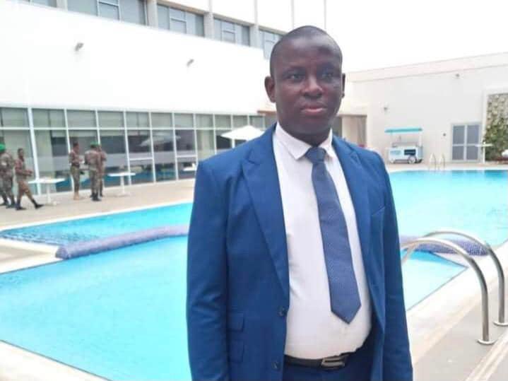 Un homme en costume bleu posé debout devant une piscine avec un bâtiment en arrière-plan et des militaires à distance