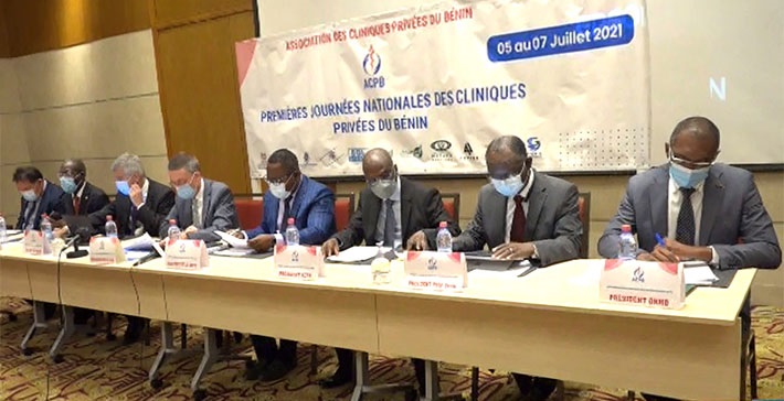 Sept hommes en costume et masques assis derrière une longue table lors d'une conférence dans une salle de réunion
