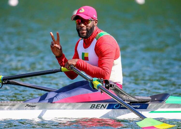 Rameur en tenue sportive rouge et blanche avec drapeau sur un bateau d’aviron faisant un signe de victoire sur l’eau