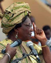 Femme portant une coiffe traditionnelle et des lunettes, parlant devant plusieurs micros lors d'un discours public