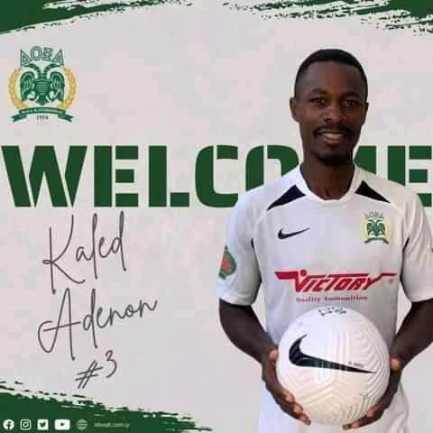 Un homme souriant en maillot de football blanc tient un ballon devant un mur blanc affichant un message de bienvenue en vert