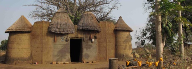 Habitation traditionnelle construite en terre avec toits de chaume coniques entourée d’arbres et d’objets colorés suspendus
