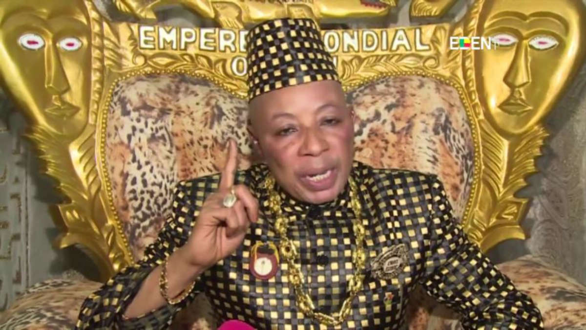 Un homme en tenue traditionnelle à motifs noirs et jaunes, assis sur un fauteuil doré orné de visages, parlant et levant un doigt