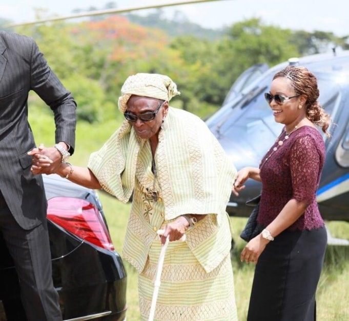 Une femme âgée en tenue traditionnelle est aidée à marcher par deux personnes près d’une voiture noire et d’un hélicoptère gris.