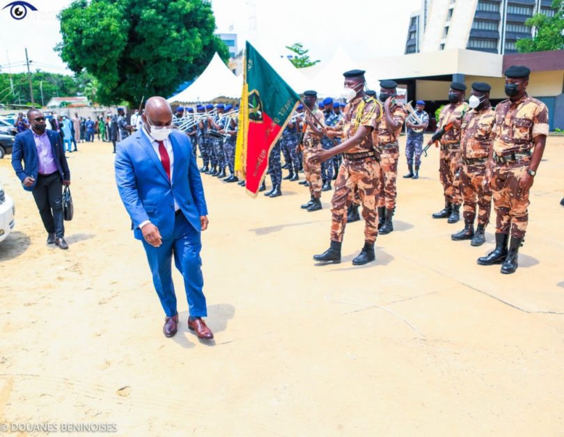 Homme en costume bleu marche devant un rang de militaires en uniforme camouflé tenant un drapeau vert rouge jaune