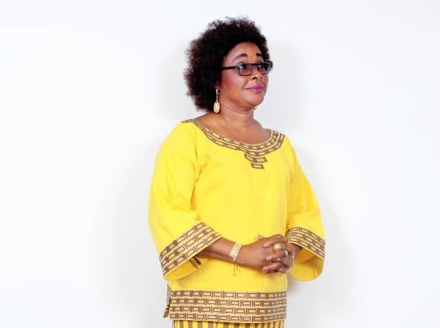 Femme debout, vêtue d’une robe jaune ornée de motifs marron, les mains jointes et regard tourné vers la droite