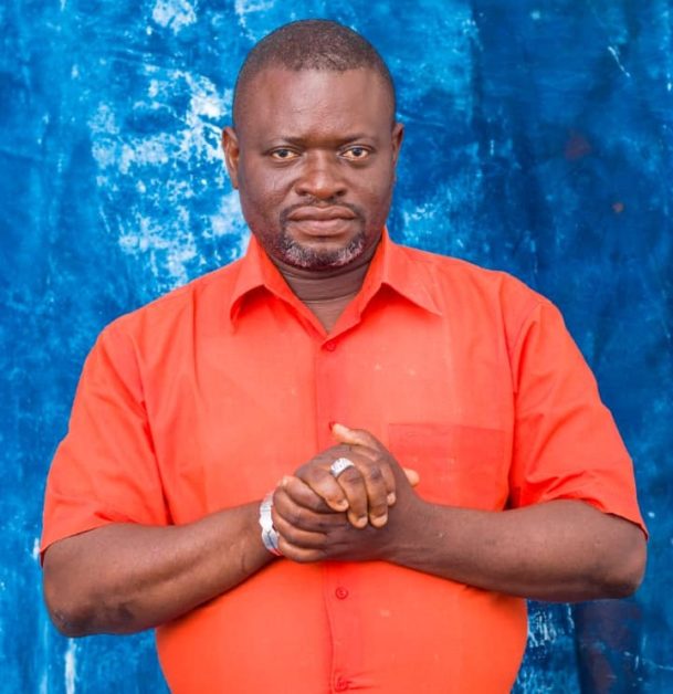 Homme portant une chemise orange se tenant devant un fond bleu avec les mains jointes au niveau de la poitrine