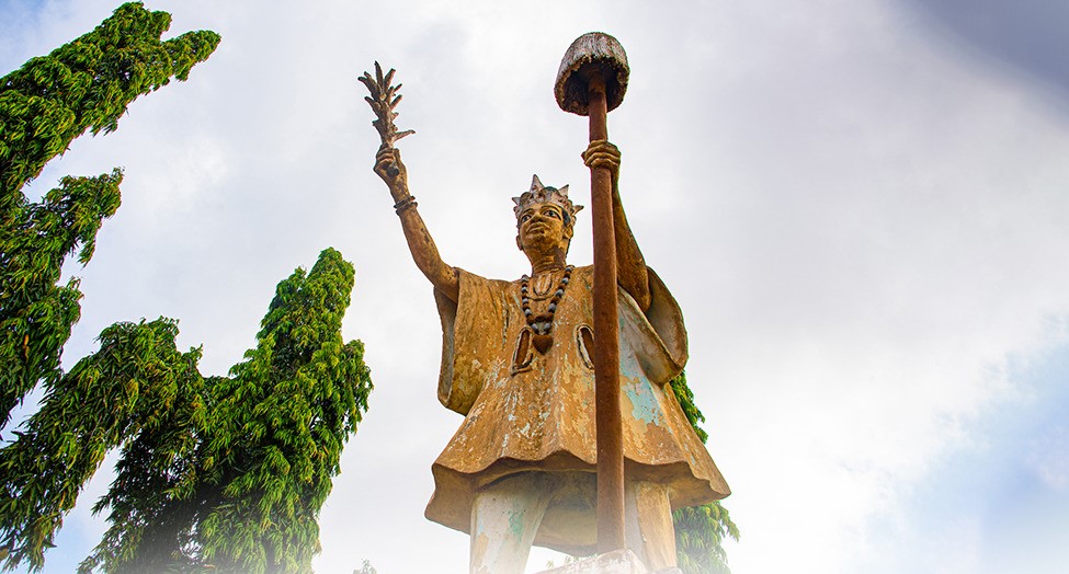 Statue d’une figure royale tenant un sceptre dans une main et un objet ornemental dans l’autre, entourée d’arbres verts