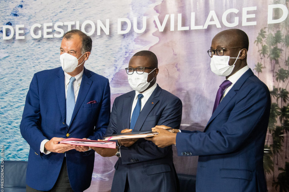 Trois hommes en costume et masque médical échangent des documents lors d’une cérémonie officielle intérieure