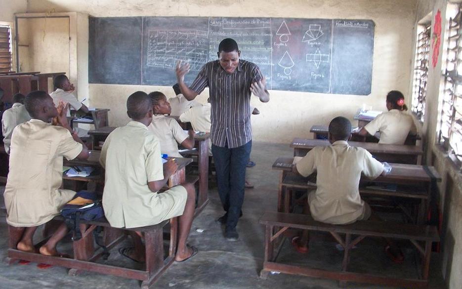 Un enseignant debout parle avec énergie à des élèves assis sur des bancs dans une salle de classe avec un tableau noir en arrière-plan