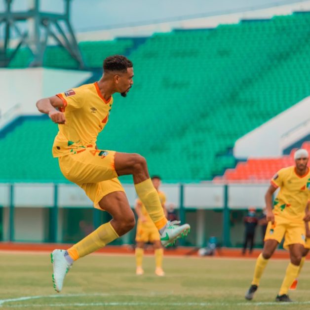 Joueur en maillot jaune en train de frapper le ballon en l'air sur un terrain de football devant des gradins verts et blancs