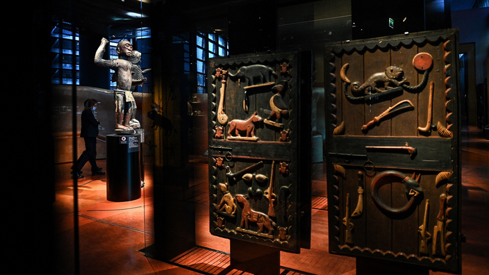 Salle d’exposition montrant des panneaux sculptés colorés et une statue disposée dans un espace tamisé avec un visiteur en arrière-plan