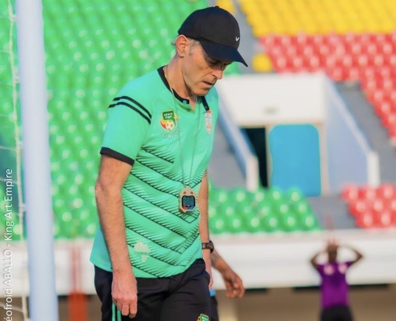 Un homme en tenue de sport et casquette noire marche sur un terrain avec des gradins multicolores en arrière-plan