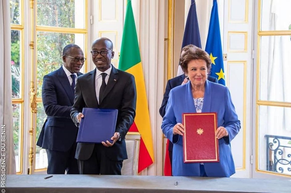 Quatre personnes en costume tenant des dossiers devant un bureau avec en arrière-plan deux drapeaux, un vert-jaune-rouge et un bleu aux étoiles jaunes