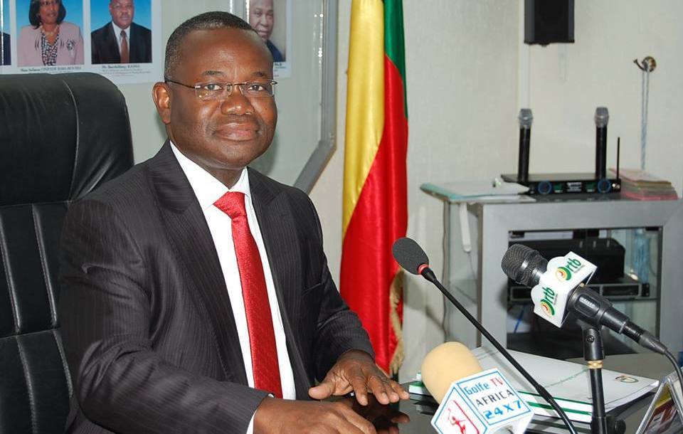 Un homme en costume sombre et cravate rouge est assis à un bureau avec plusieurs micros devant lui et un drapeau aux couleurs vert, jaune, rouge à l'arrière-pla
