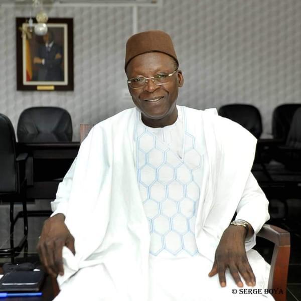 Homme souriant en tenue traditionnelle blanche et chapeau assis dans un bureau avec un portrait au mur derrière lui