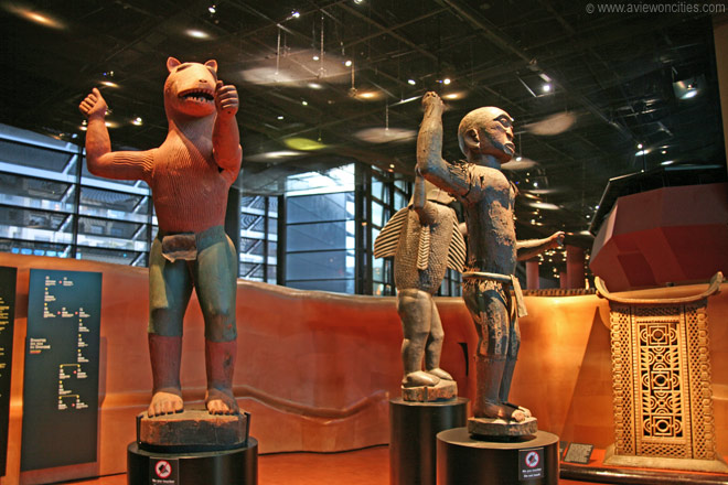 Trois grandes statues traditionnelles en bois coloré exposées dans une salle aux murs ocres et au plafond sombre lumineux