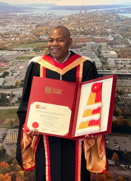 Homme en toge de docteur honoraire tenant un diplôme et une écharpe rouge et jaune devant une vue aérienne urbaine
