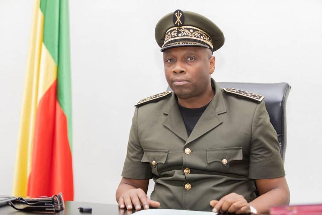 Un homme en uniforme militaire assis devant un bureau avec un drapeau tricolore en arrière-plan