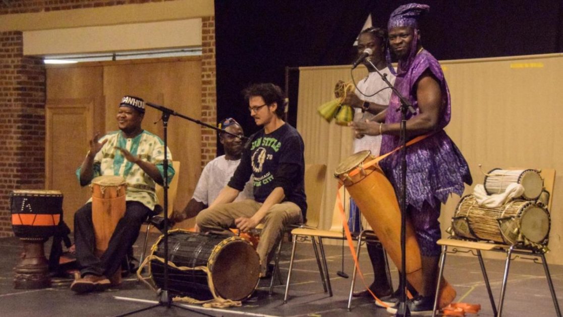 Quatre musiciens jouent des percussions sur scène avec des tenues traditionnelles et décontractées dans une salle.