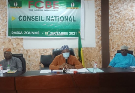 Trois hommes assis derrière une table lors d’une réunion sous une bannière verte indiquant un conseil national