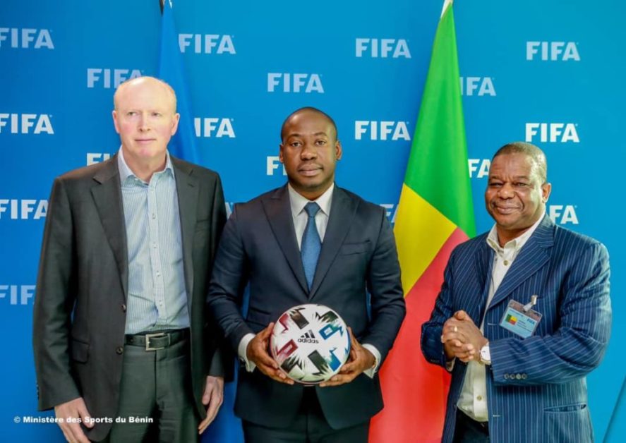 Trois hommes en costume posent devant un drapeau tricolore avec un ballon de football devant un fond bleu marqué FIFA